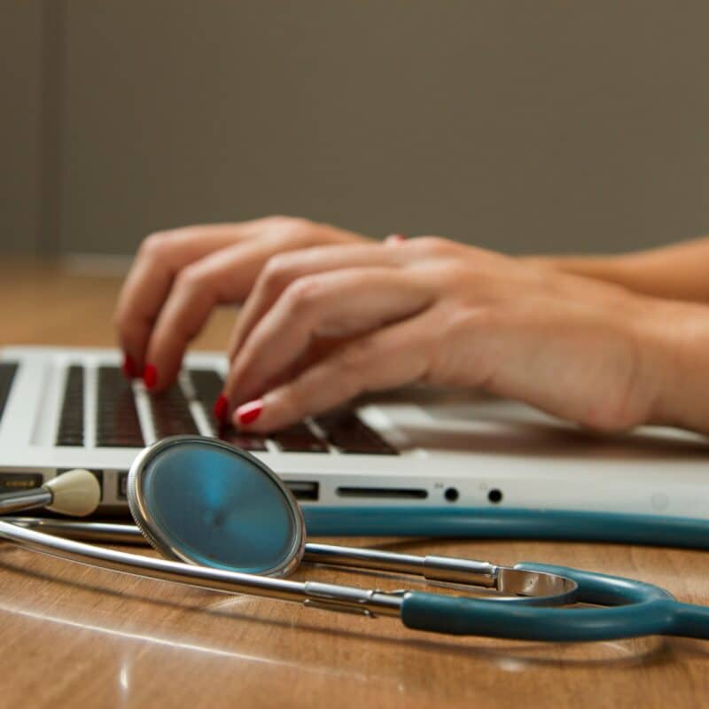 A stethoscope beside a person typing on a laptop, emphasizing the importance of seeking guidance from health professionals.