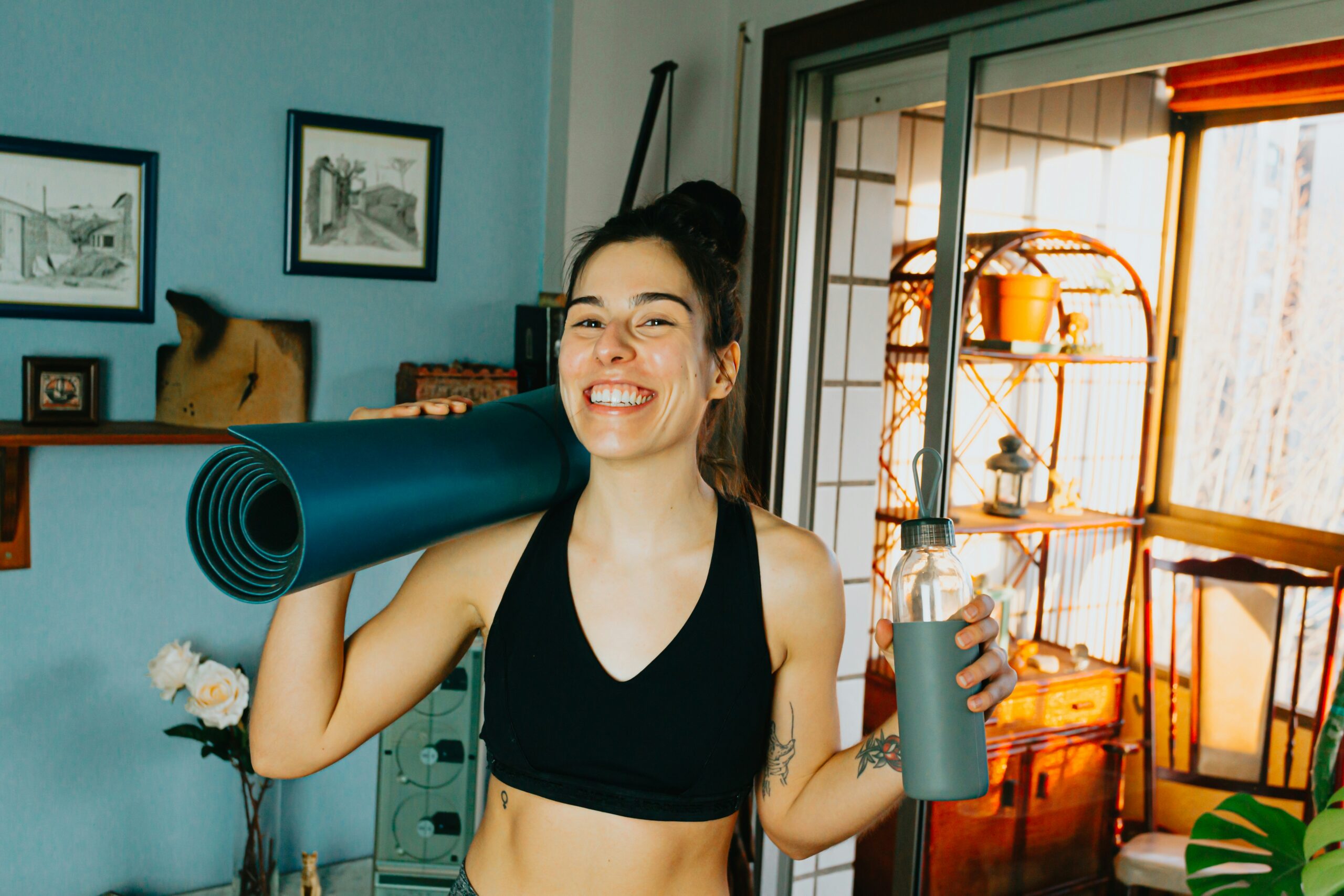 A person smiling while holding a yoga mat over one shoulder and a water bottle in the other hand, ready to practice yoga, reflecting an active and healthy lifestyle.