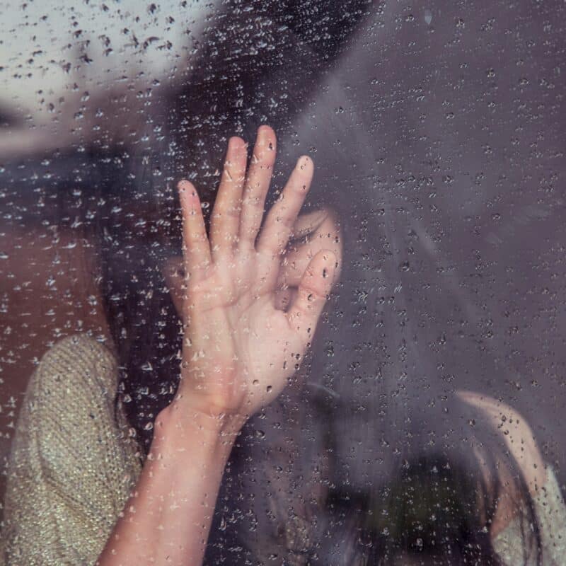 A person behind a rain-covered window reaches out with their hand, symbolizing the emotional isolation and challenges anxiety can add to managing endometriosis.