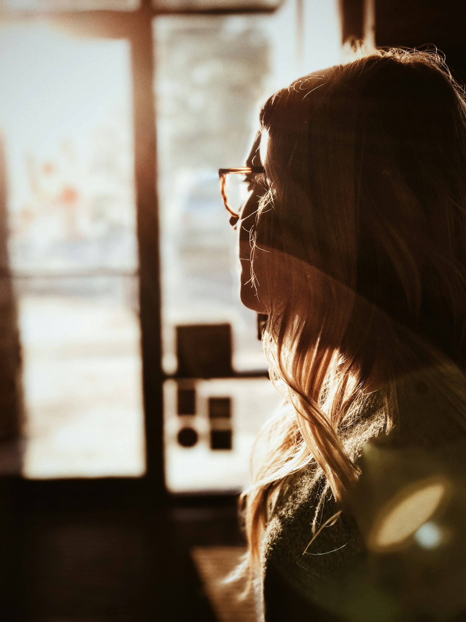 A woman with glasses gazing thoughtfully out of a window, bathed in warm sunlight, reflecting a sense of contemplation about stress related to endometriosis.