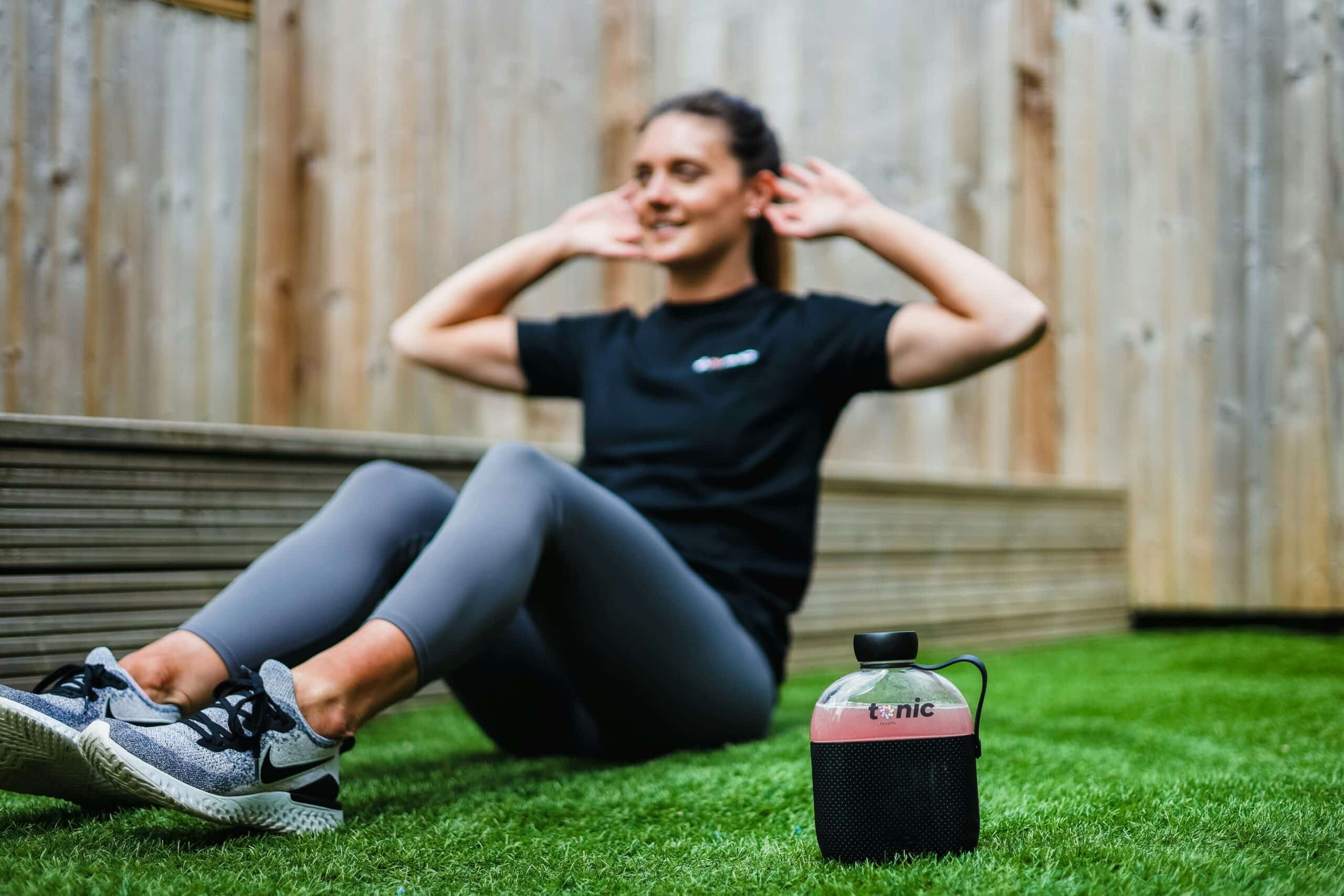 Woman performing core exercises on grass, emphasizing physical activity and wellness, with a water bottle labeled "tonic" nearby, illustrating an active lifestyle relevant to pelvic floor physical therapy and integrative endometriosis treatment.