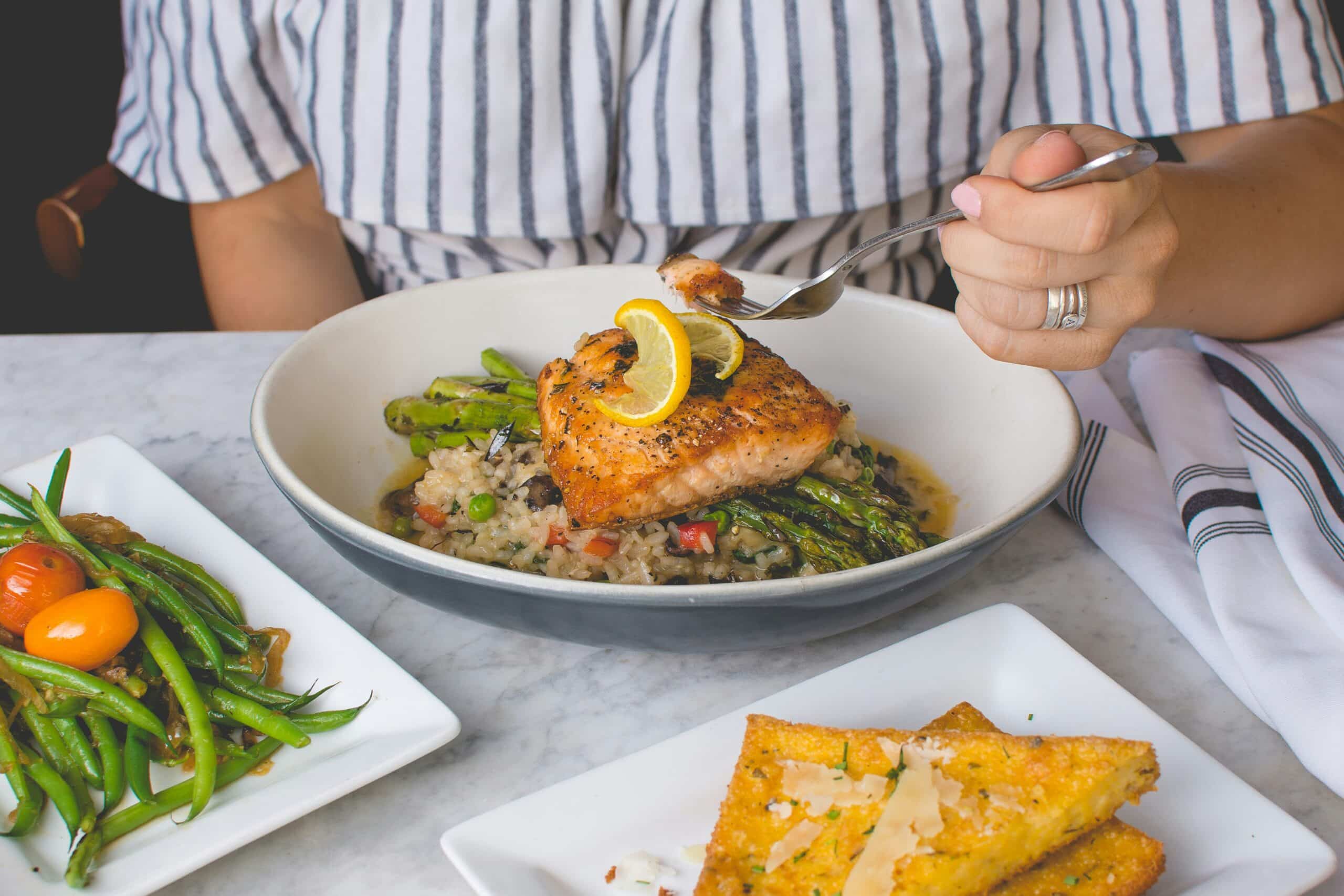 A person enjoying a meal featuring grilled salmon with vegetables and risotto, highlighting the safety and considerations of omega-3 fatty acid consumption in a balanced diet.