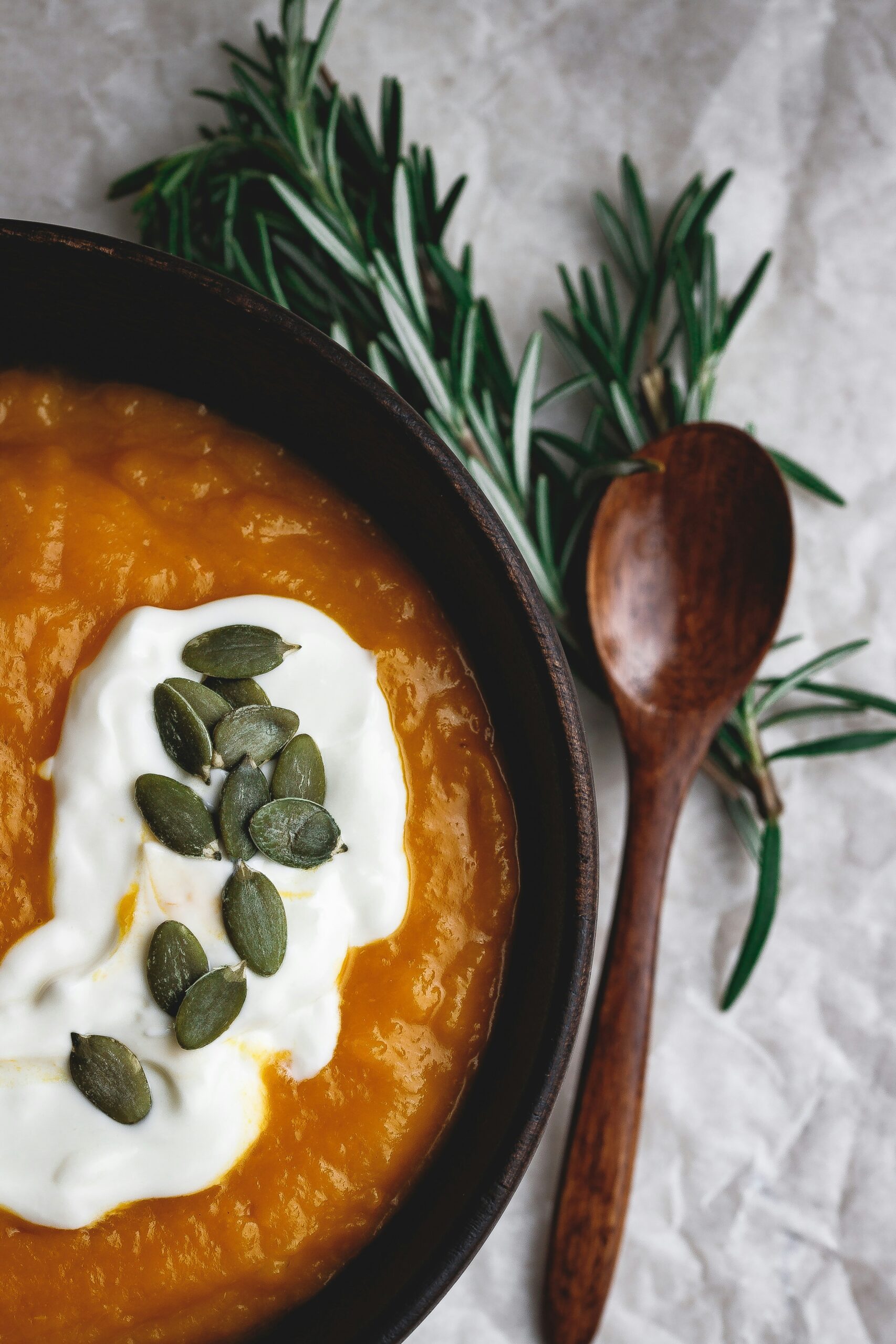 A bowl of creamy pumpkin soup topped with yogurt and pumpkin seeds, placed next to fresh rosemary and a wooden spoon, representing a nutrient-rich meal for holistic nutrition in managing endometriosis. - Rachael Haverland Site A bowl of creamy pumpkin soup topped with yogurt and pumpkin seeds, placed next to fresh rosemary and a wooden spoon, representing a nutrient-rich meal for holistic nutrition in managing endometriosis.