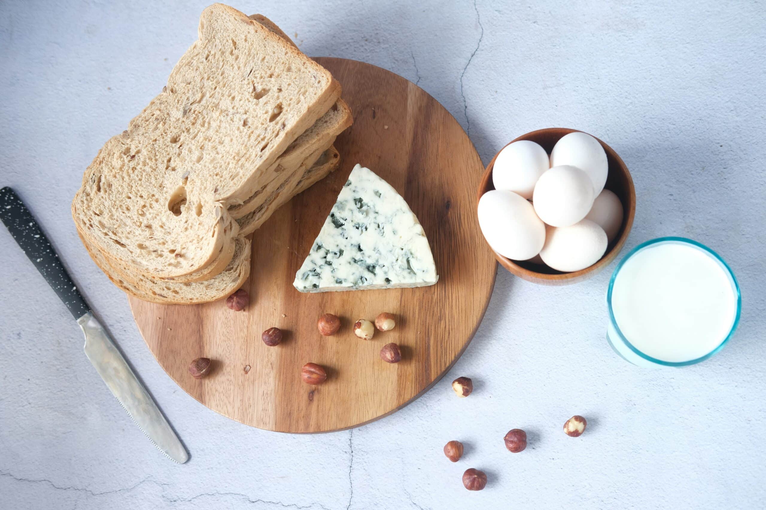 A wooden board with bread, blue cheese, eggs, hazelnuts, and a glass of milk, representing the evaluation of dairy products and their components in nutrition and dietary choices.