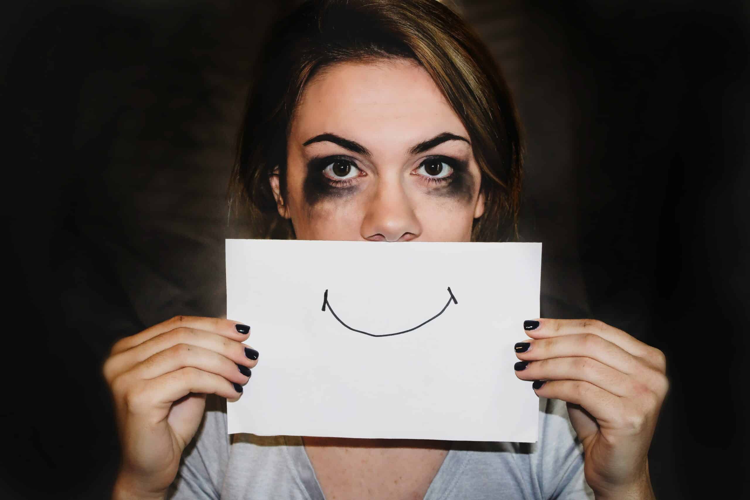 Woman with smudged makeup holds a paper smile over her mouth, symbolizing hidden mood swings often experienced with endometriosis. - Rachael Haverland Site Woman with smudged makeup holds a paper smile over her mouth, symbolizing hidden mood swings often experienced with endometriosis.
