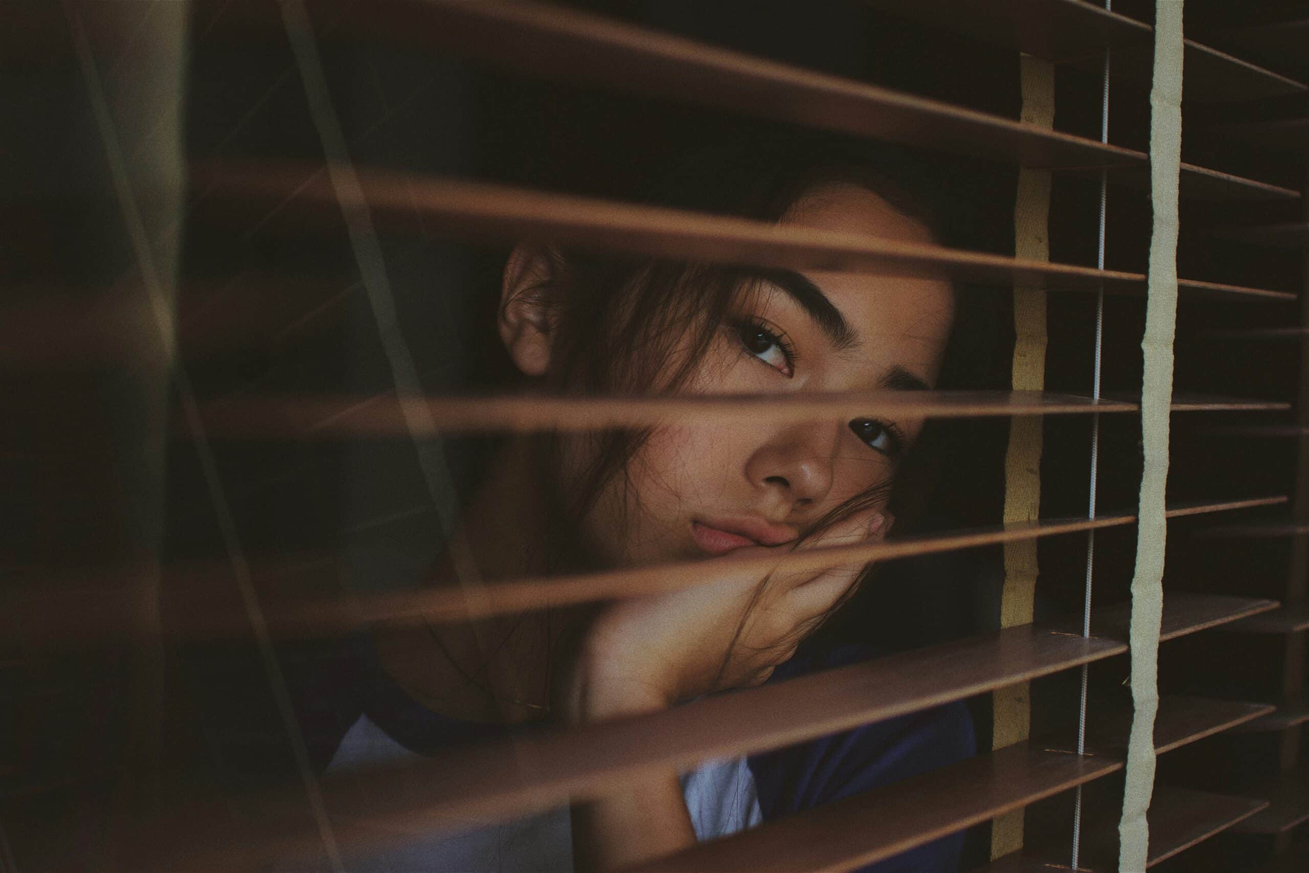 A woman rests her face on her hand, gazing pensively through wooden blinds, symbolizing the emotional depth behind lifestyle changes supporting endometriosis fertility. - Rachael Haverland Site A woman rests her face on her hand, gazing pensively through wooden blinds, symbolizing the emotional depth behind lifestyle changes supporting endometriosis fertility.