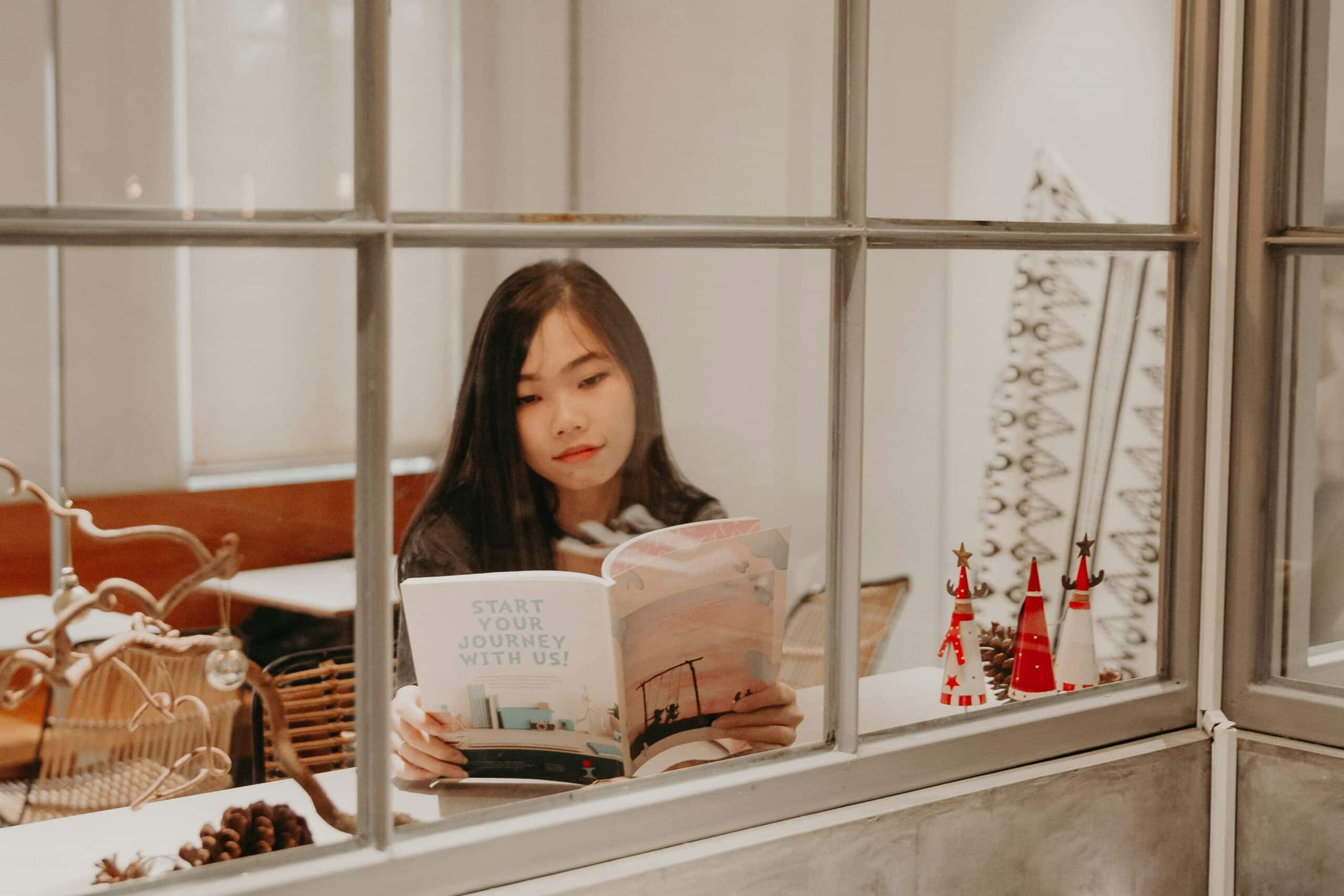 A young woman reads a magazine titled "Start Your Journey With Us" by a window, symbolizing the importance of exploring additional resources for guidance and support. - Rachael Haverland Site A young woman reads a magazine titled "Start Your Journey With Us" by a window, symbolizing the importance of exploring additional resources for guidance and support.