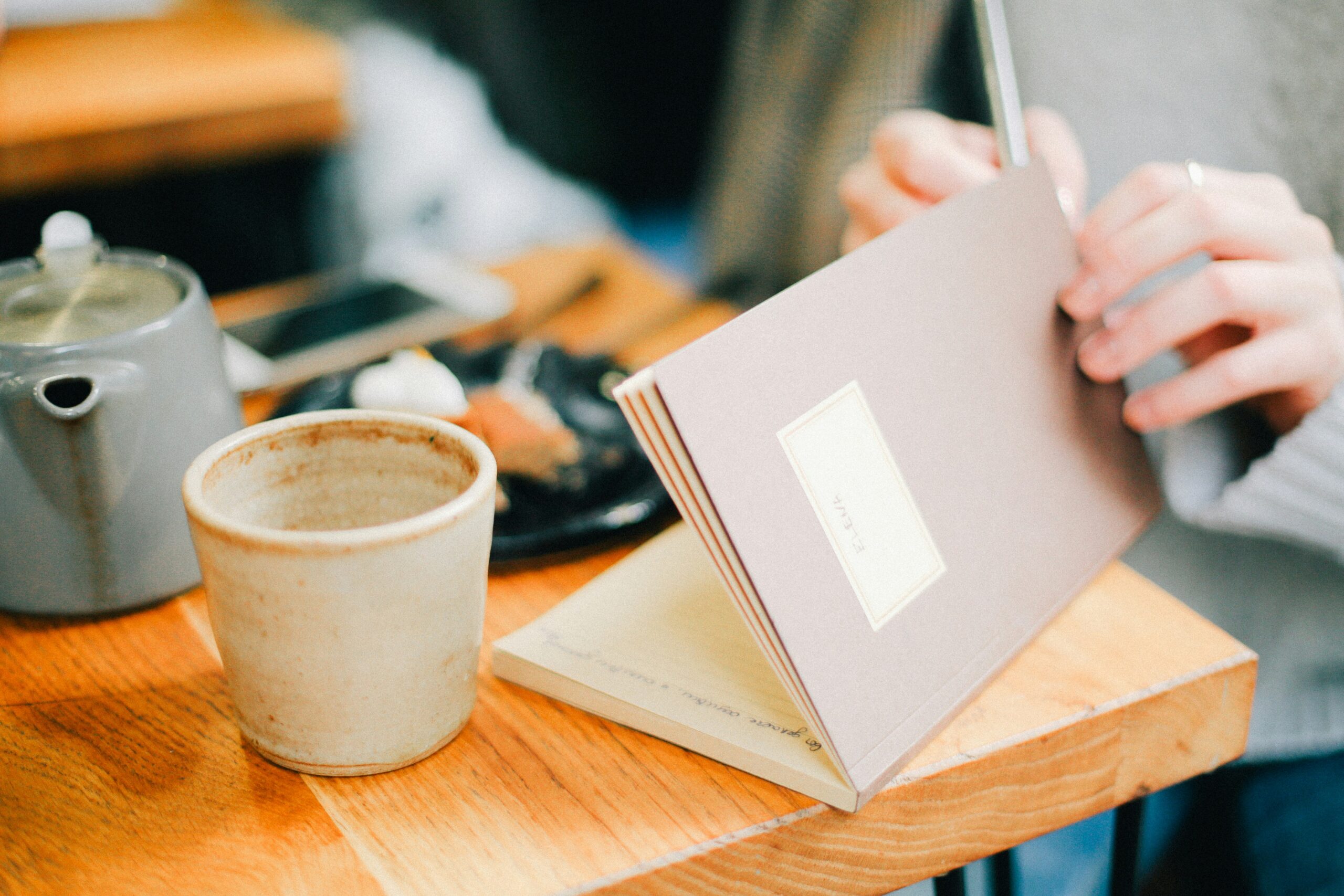 Person writing in a notebook at a café table with a cup of tea and a plate of food, emphasizing mindfulness and symptom tracking for digestive health management.