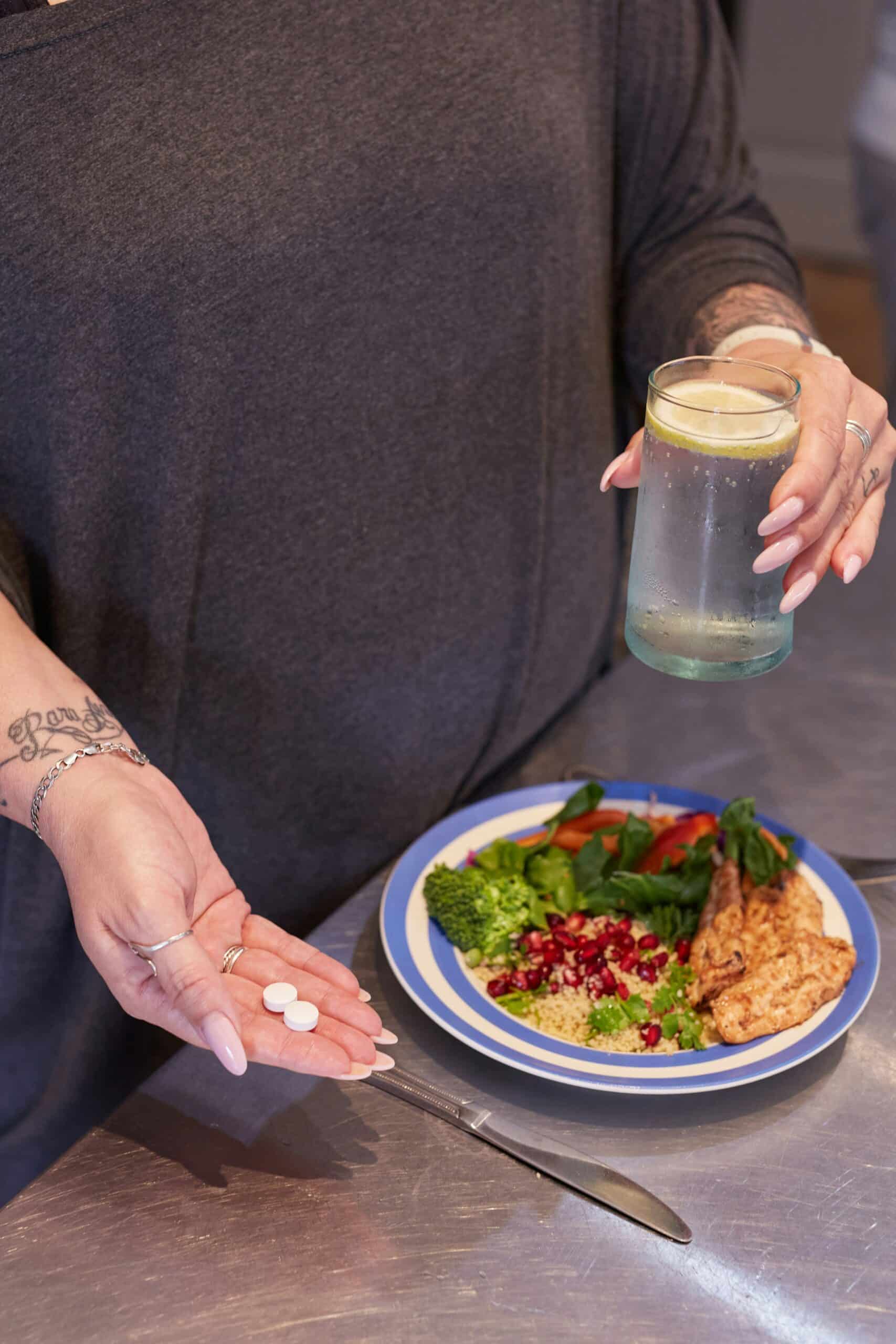 Person holding tablets and a glass of lemon water near a healthy meal, emphasizing conscious food choices to manage endometriosis symptoms. - Rachael Haverland Site Person holding tablets and a glass of lemon water near a healthy meal, emphasizing conscious food choices to manage endometriosis symptoms.