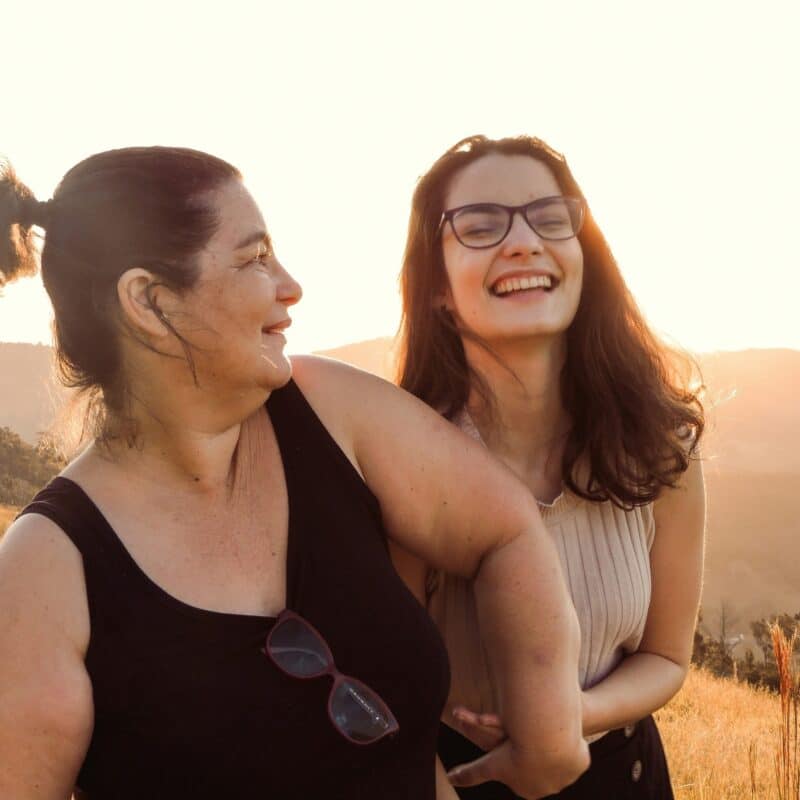 Two women smiling and laughing together in a sunlit outdoor setting, representing support and connection, relevant to discussions on family medical history in endometriosis care.
