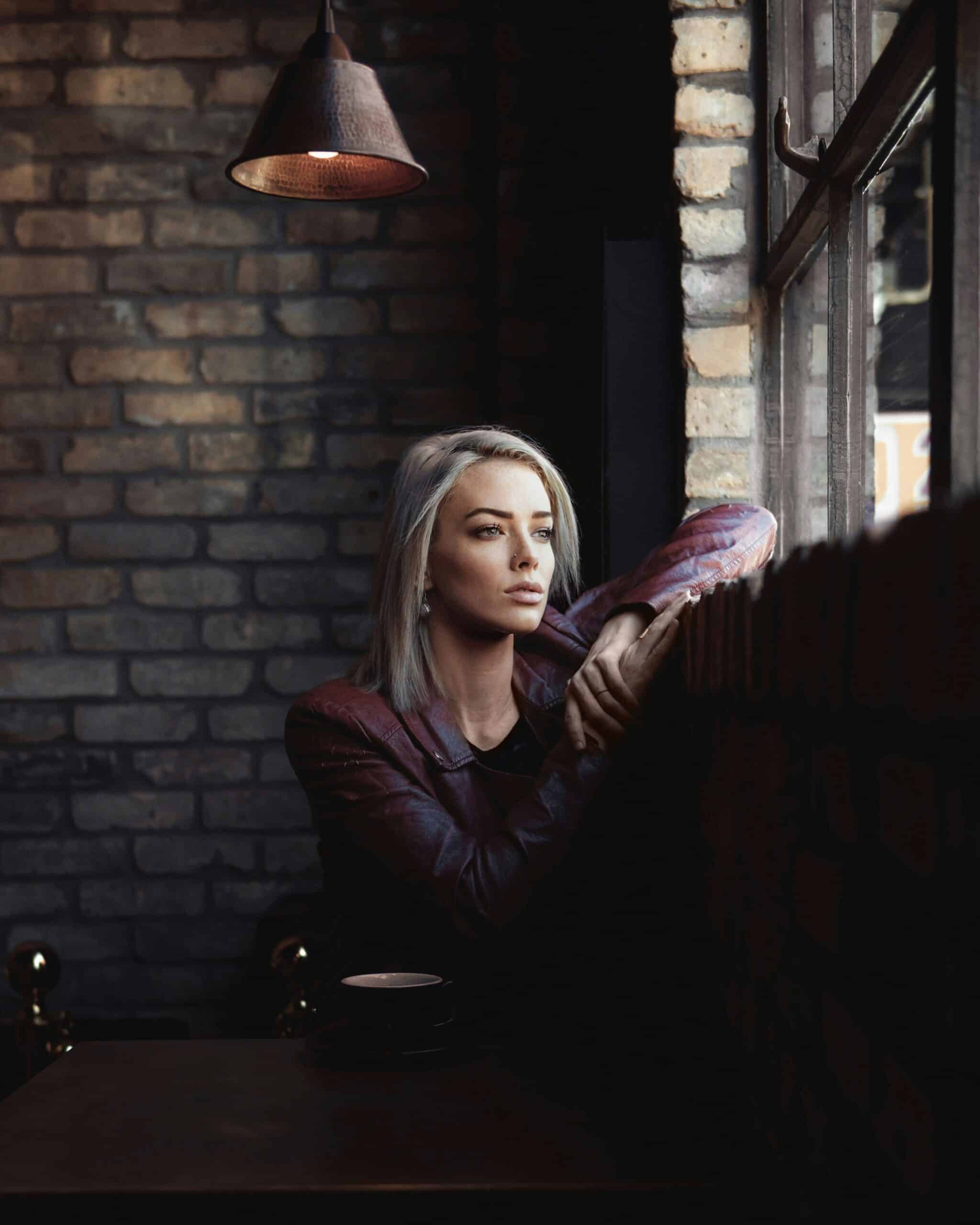 Woman with short blonde hair sitting by a window in a cozy café, looking contemplative, reflecting the emotional toll of chronic conditions like endometriosis.