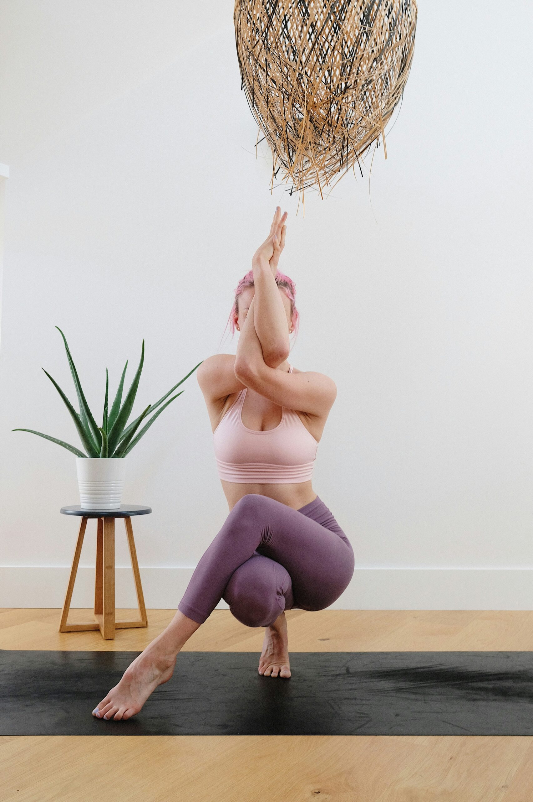 Woman performing a yoga pose on a black mat, demonstrating flexibility and strength, with a potted plant and wooden stool in a bright indoor setting, relevant to pelvic floor exercises and relaxation techniques for endometriosis relief.