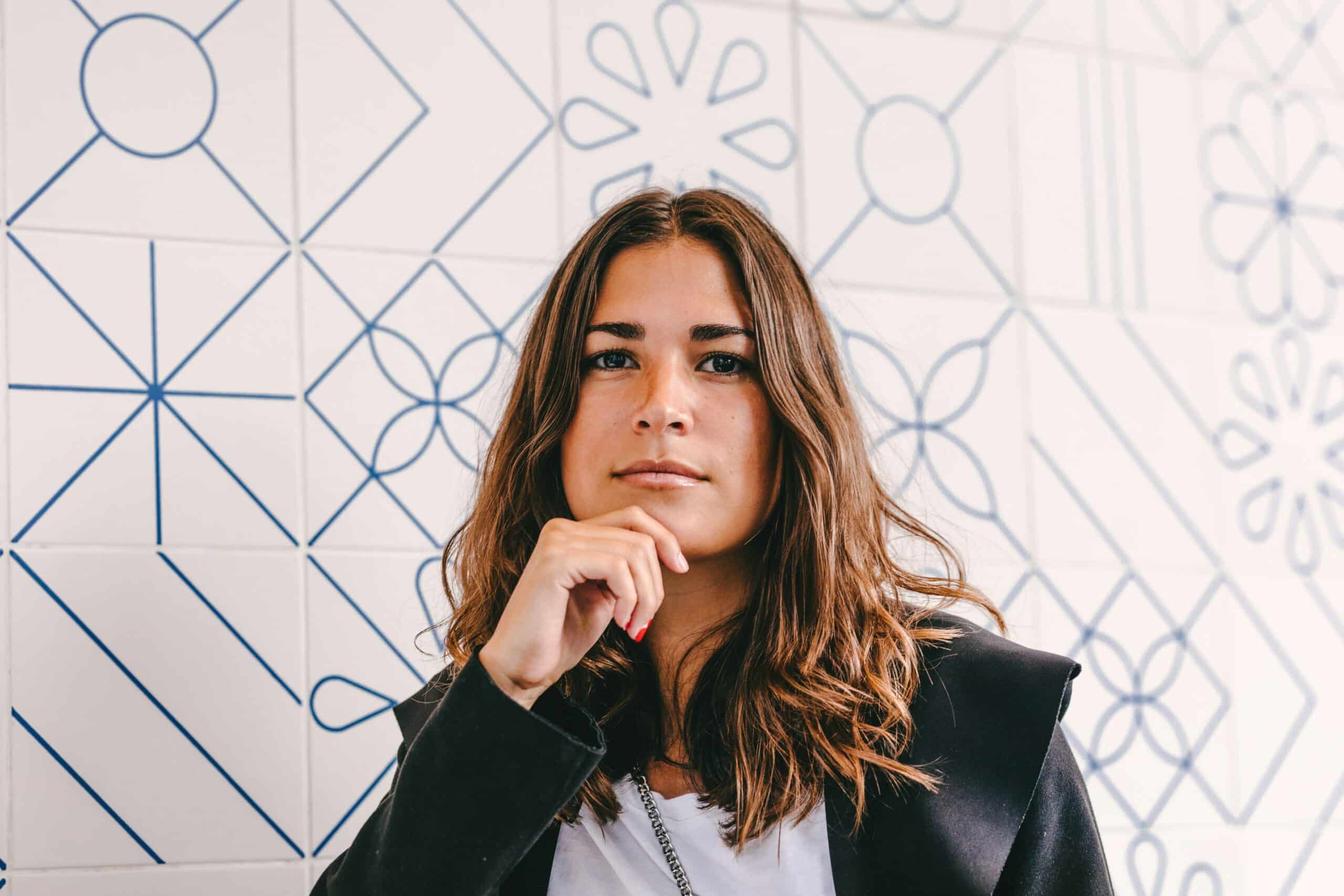 Young woman with long brown hair, thoughtfully resting her chin on her hand, against a patterned blue and white tiled background, conveying a sense of contemplation related to pelvic floor therapy and endometriosis management.