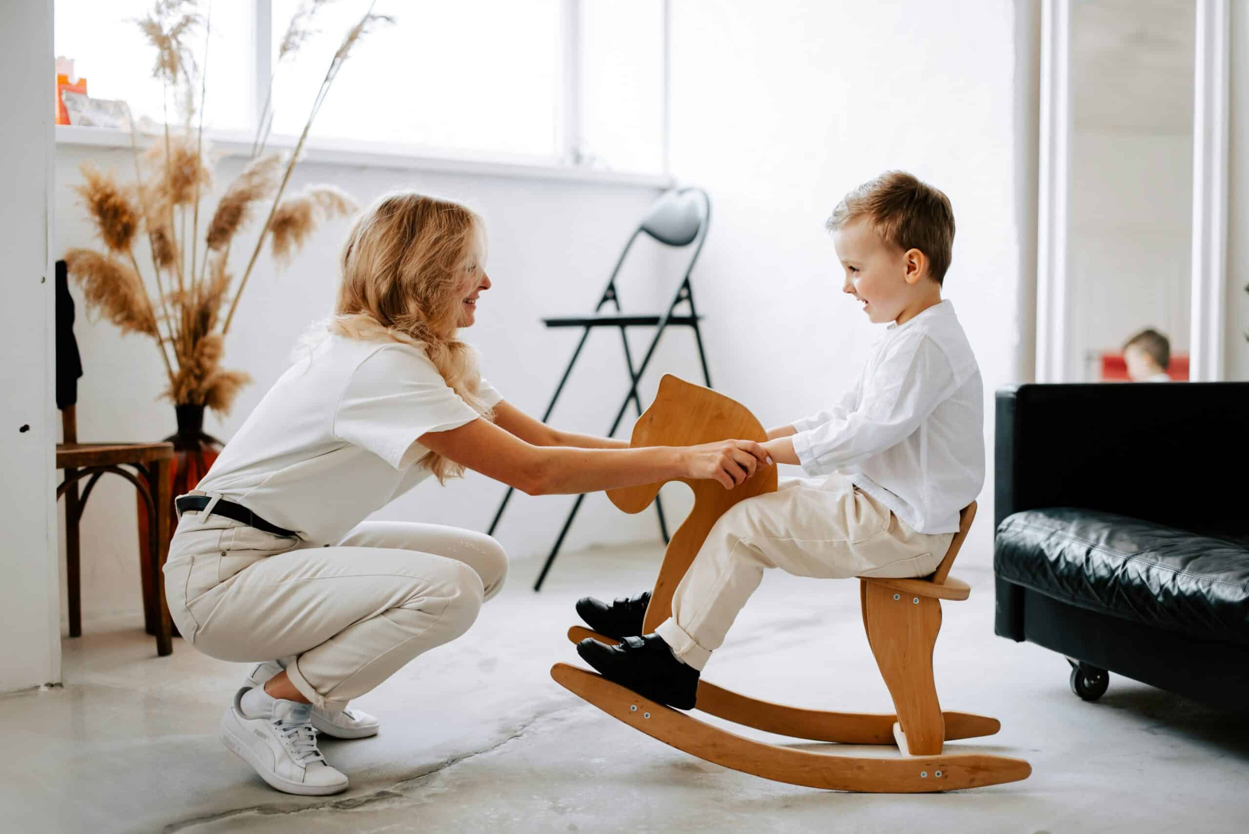 Woman engaging with a child on a wooden rocking horse in a bright, modern space, illustrating playful interaction and nurturing environment.