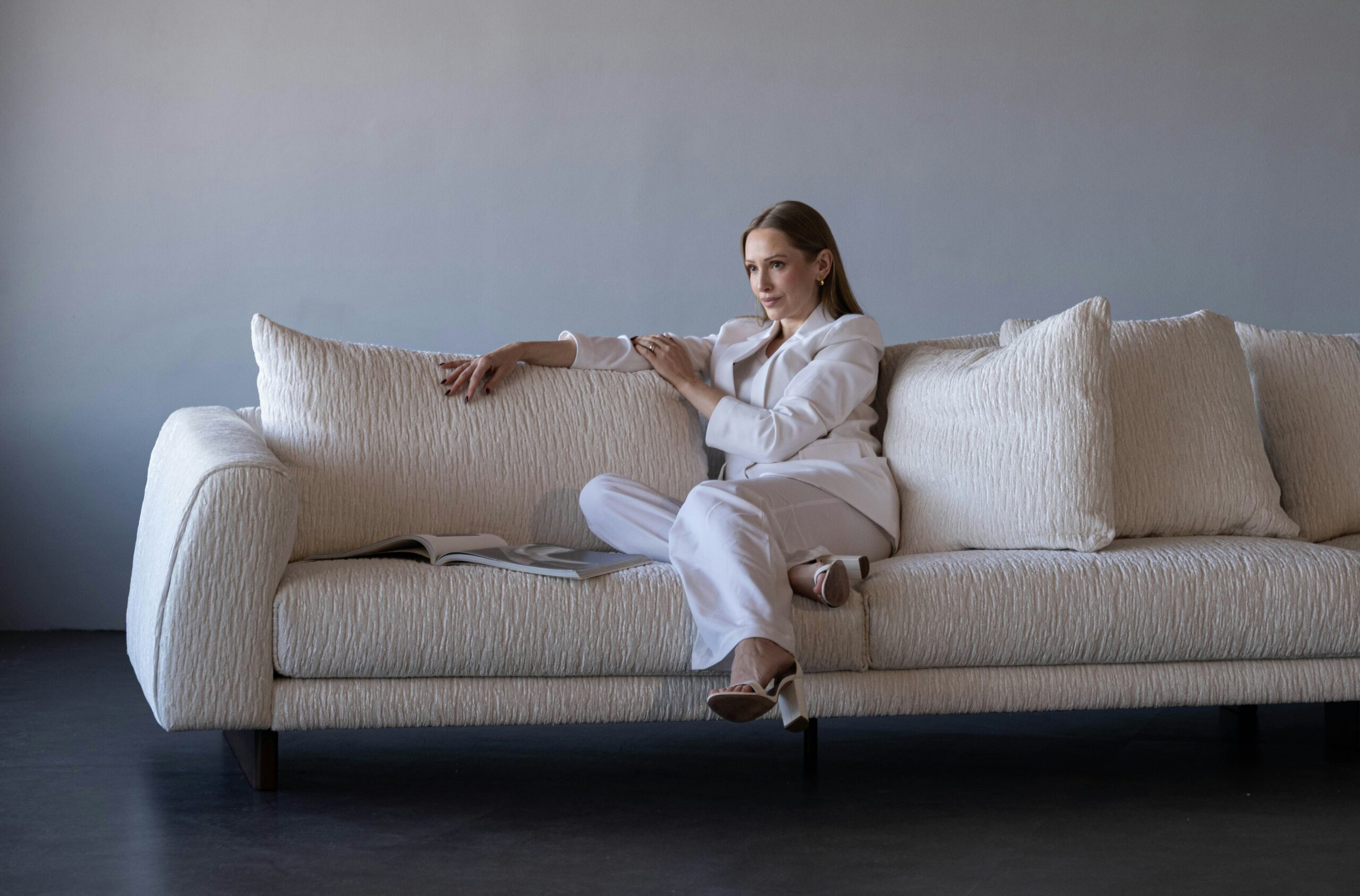Woman in a white suit seated on a textured beige sofa, embodying a calm and confident demeanor, with a magazine resting beside her, reflecting a serene and modern interior setting.