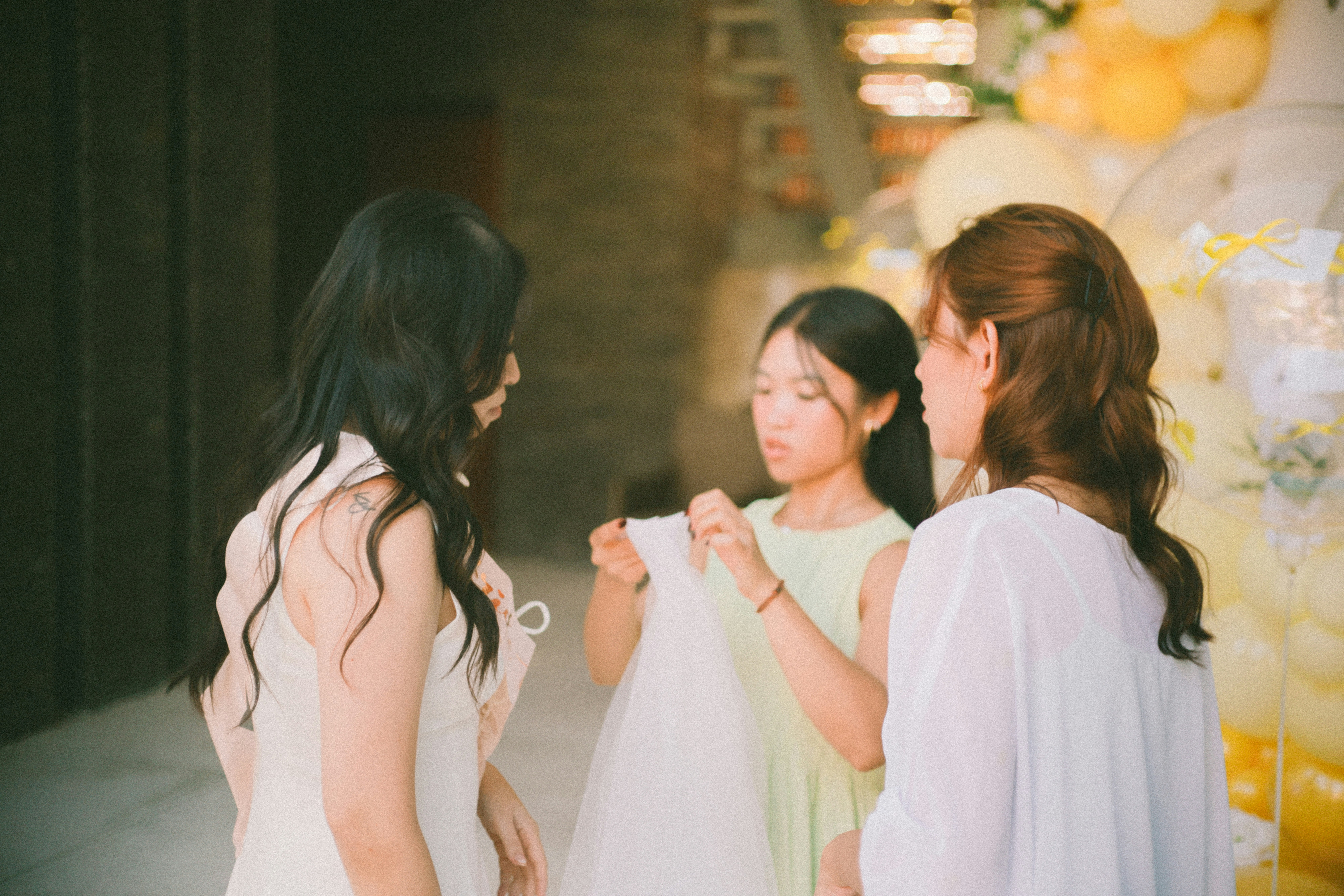 Three women discussing and handling fabric in a celebratory setting, surrounded by decorative balloons, emphasizing community and support.