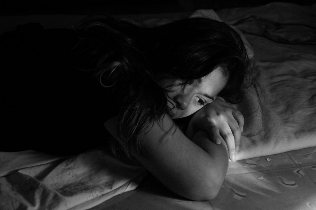 A person lying on a bed in low light, head turned to the side and hands resting under the chin.