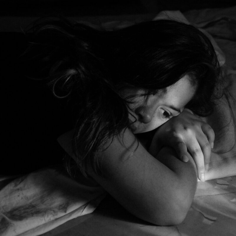A person lying on a bed in low light, head turned to the side and hands resting under the chin.